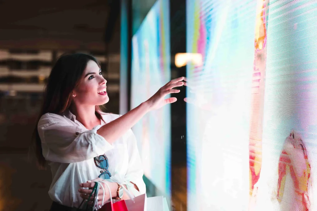 Publicidad Local | Campañas Efectivas para tu Negocio 2 Joven mujer con bolsas de compra tocando una pantalla digital interactiva, sonriendo, simbolizando la experiencia de compra atractiva y la interacción con la publicidad digital en negocios locales de Zaragoza.